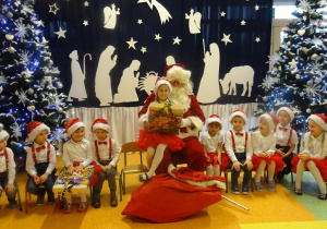 Dziewczynka siedzi na kolanach Mikołaja, ogląda prezent. Wokół siedzi grupa dzieci.